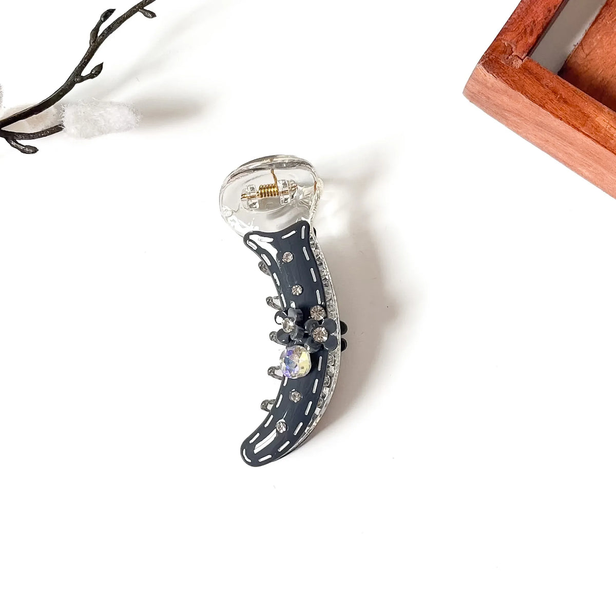 A crescent-shaped claw banana clip with a navy blue faux-suede body, clear rhinestone trim, and a large iridescent purple crystal at the center, displayed on a white surface with decorative props.