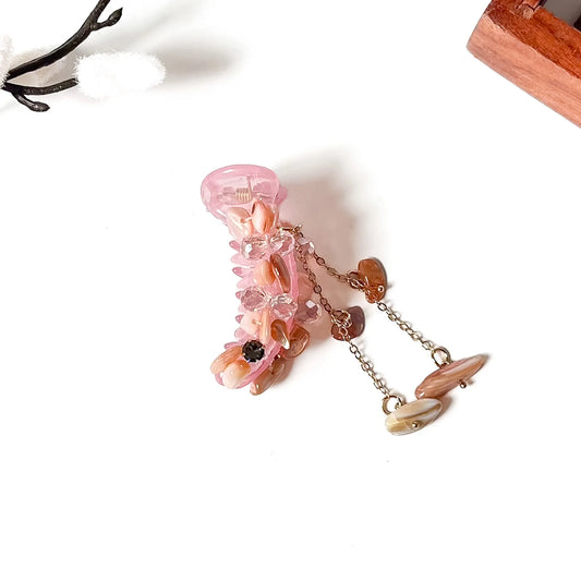 A pink translucent banana clip with embedded natural stones and dangling chain tassels, photographed on a clean white background with small decorative props.