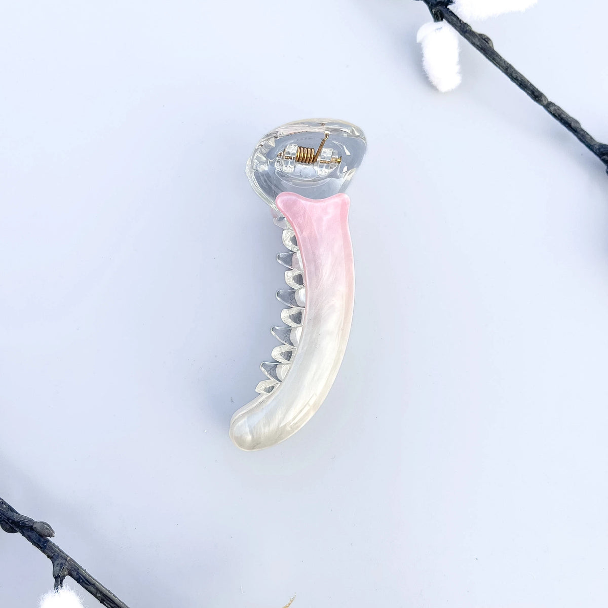 A curved banana hair clip with a translucent clear plastic body and a glossy pink inner lining, photographed on a white surface with an open book and wooden object in the background.