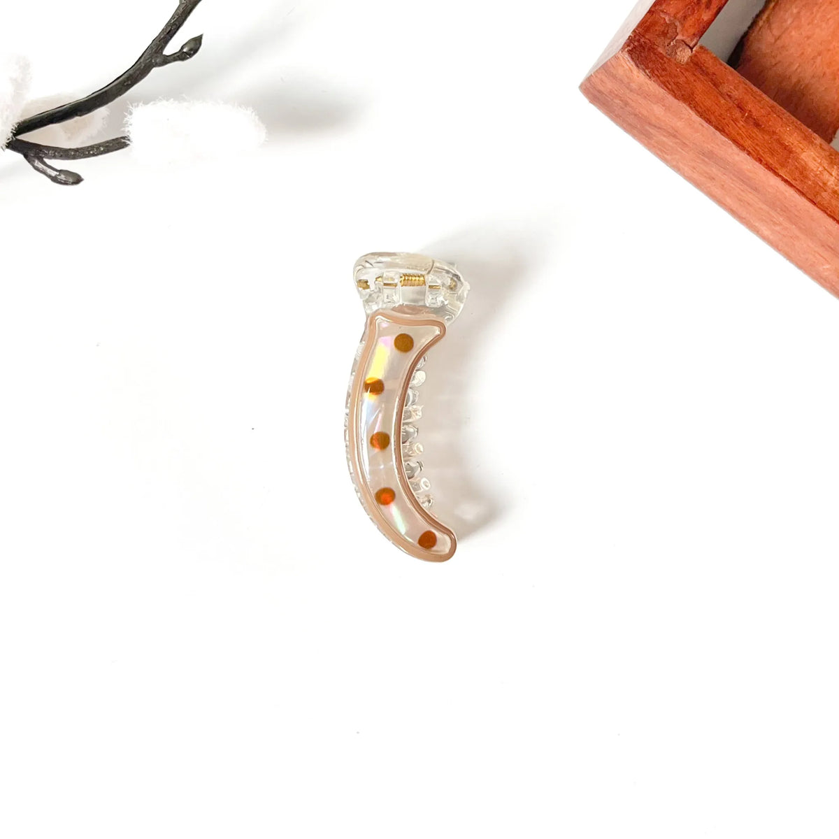 A clear crescent-shaped mini banana claw clip with amber-brown dots and a small gold accent, displayed on a white surface with decorative props.