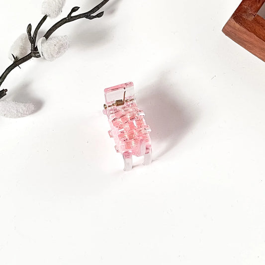 A small pink translucent small mini catch clip with a gold metal spring, photographed on a white background with decorative branches and a wooden corner visible.