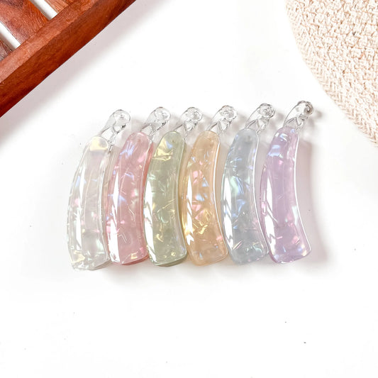 Five iridescent crescent-shaped lock banana clip with clear loops, displayed in a row on a white surface with a wooden and woven prop in the background.