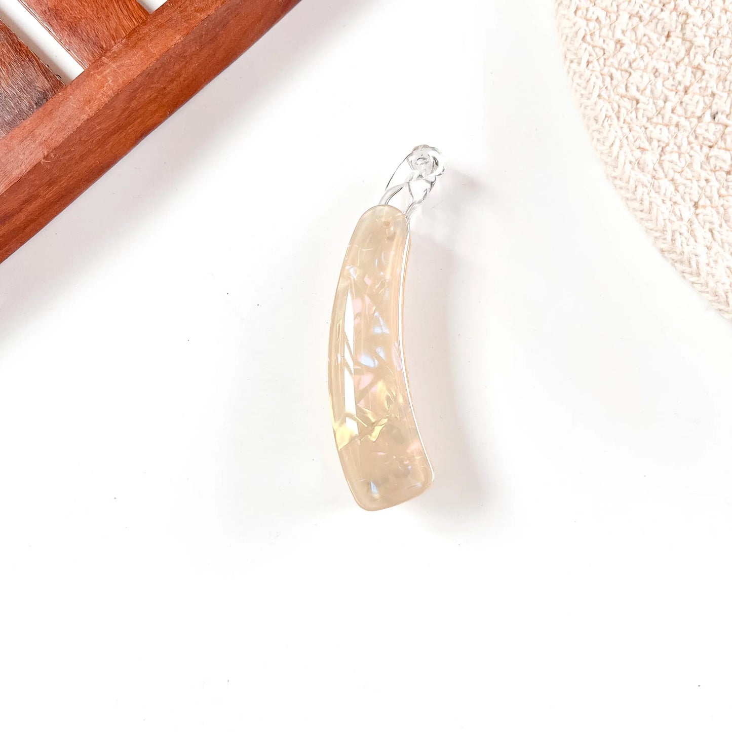 A curved, translucent pale gold lock banana clip with a small silver bail, photographed on a white surface with partial wooden and woven props at the edges.