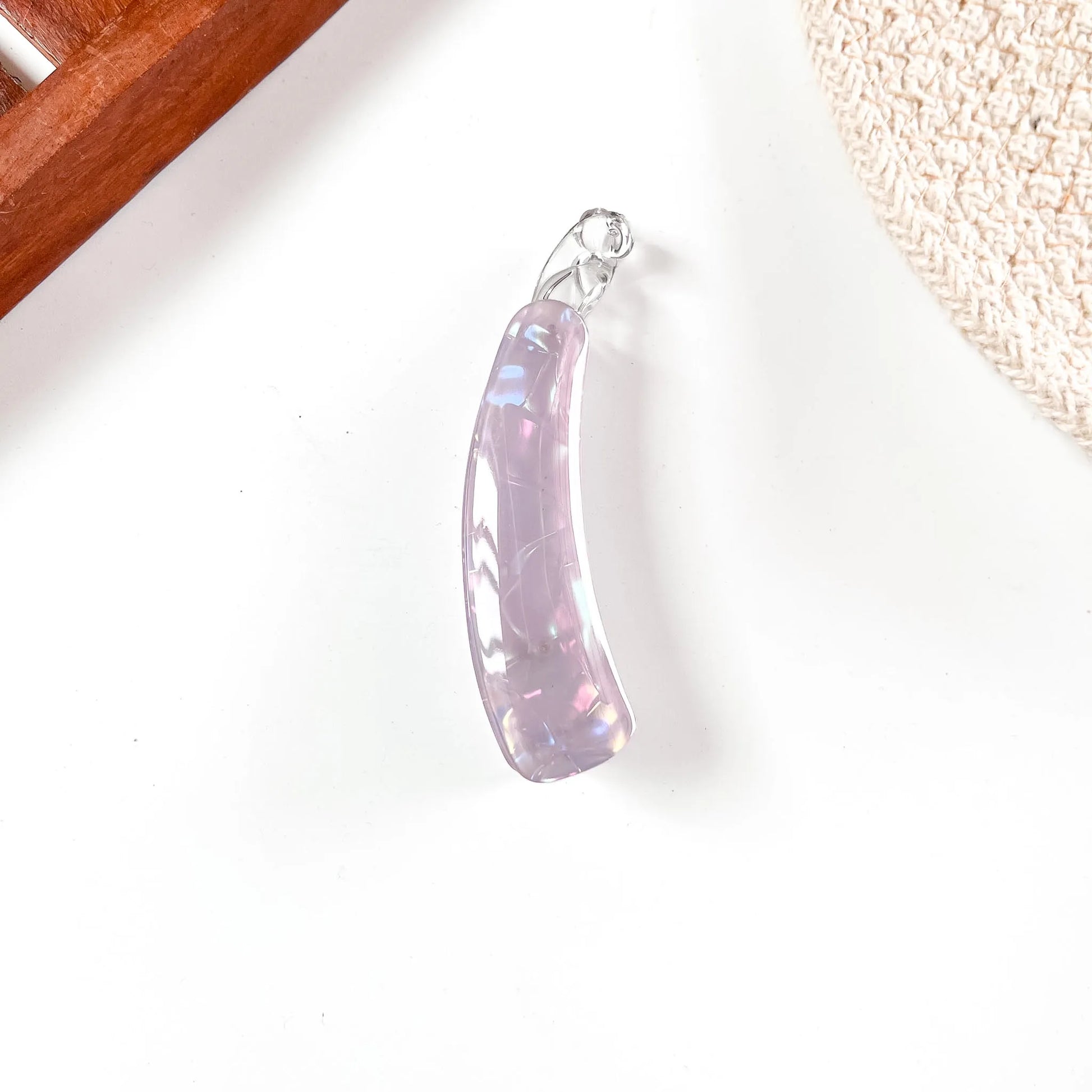 A curved iridescent lavender banana clip with a silver bail, resting on a white surface with decorative props in the corners.