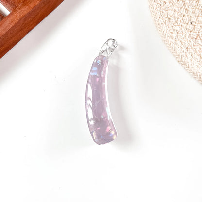 A curved iridescent lavender banana clip with a silver bail, resting on a white surface with decorative props in the corners.