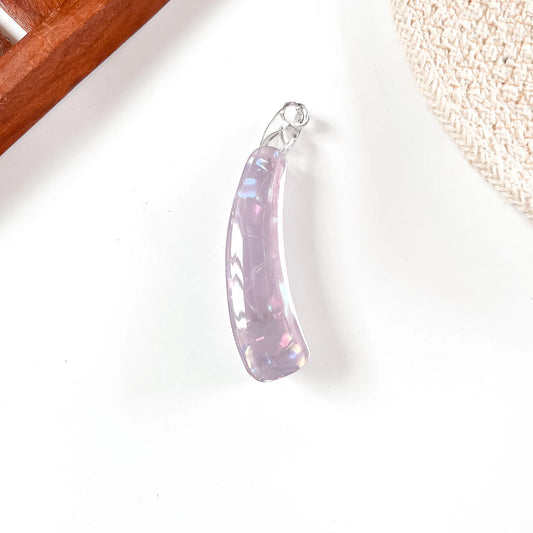 A curved iridescent lavender banana clip with a silver bail, resting on a white surface with decorative props in the corners.