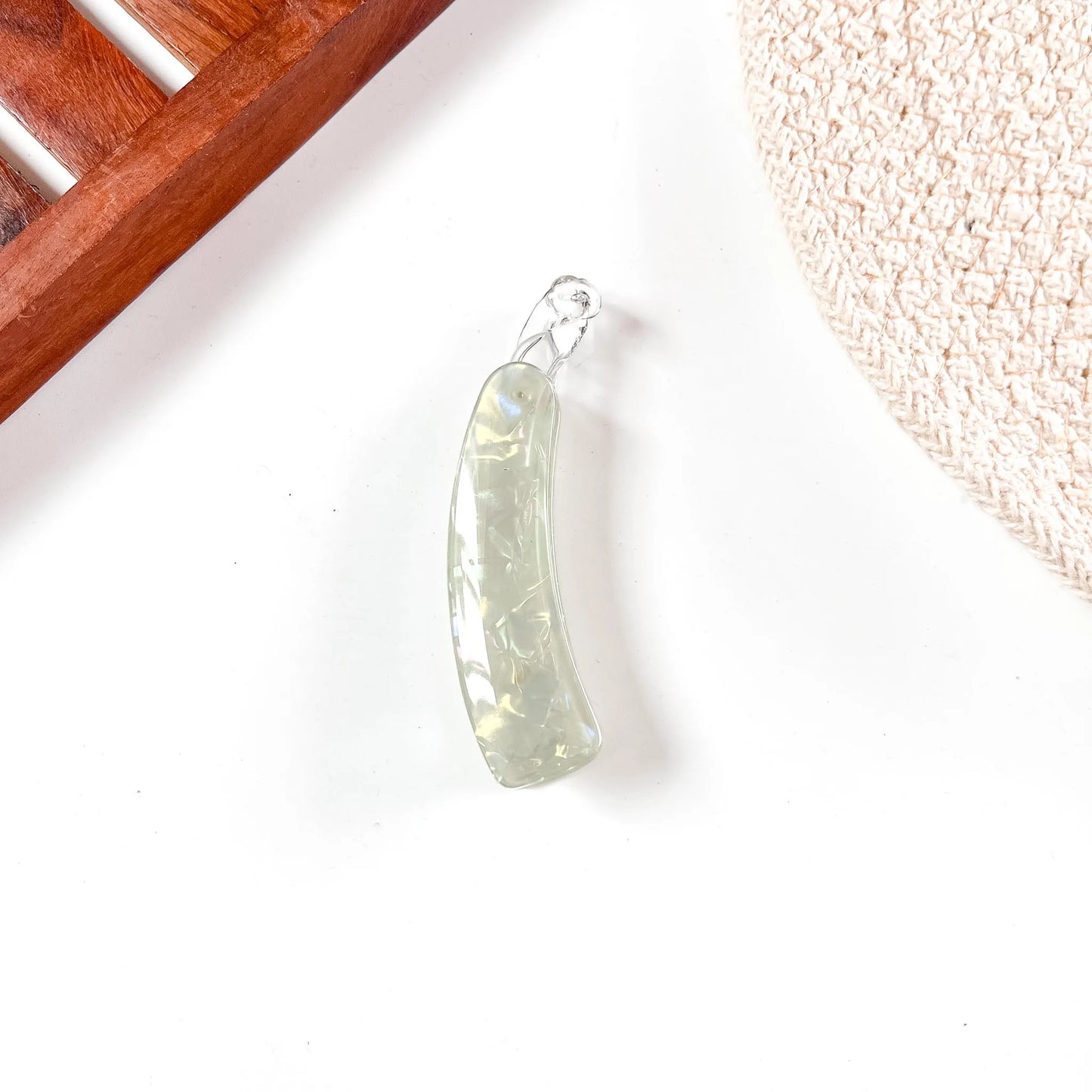 A curved, translucent pale green lock banana clip with a silver-toned metal loop at the top, resting on a white surface with decorative props in the corners.