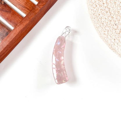 A curved, polished pink stone lock banana clip with a clear glass or acrylic loop at the top, displayed on a white surface with a wooden tray corner and a woven textile in the background.