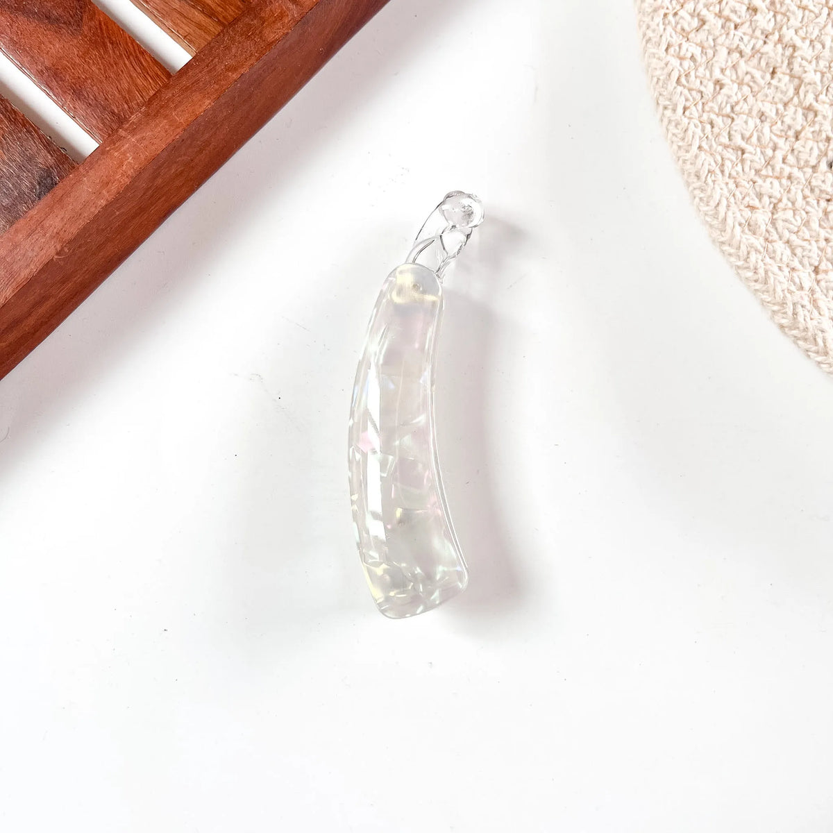 A clear, faceted glass or acrylic lock banana clip shaped like a curved teardrop or leaf, shown on a white surface with a wooden slat and beige textured fabric in the background.