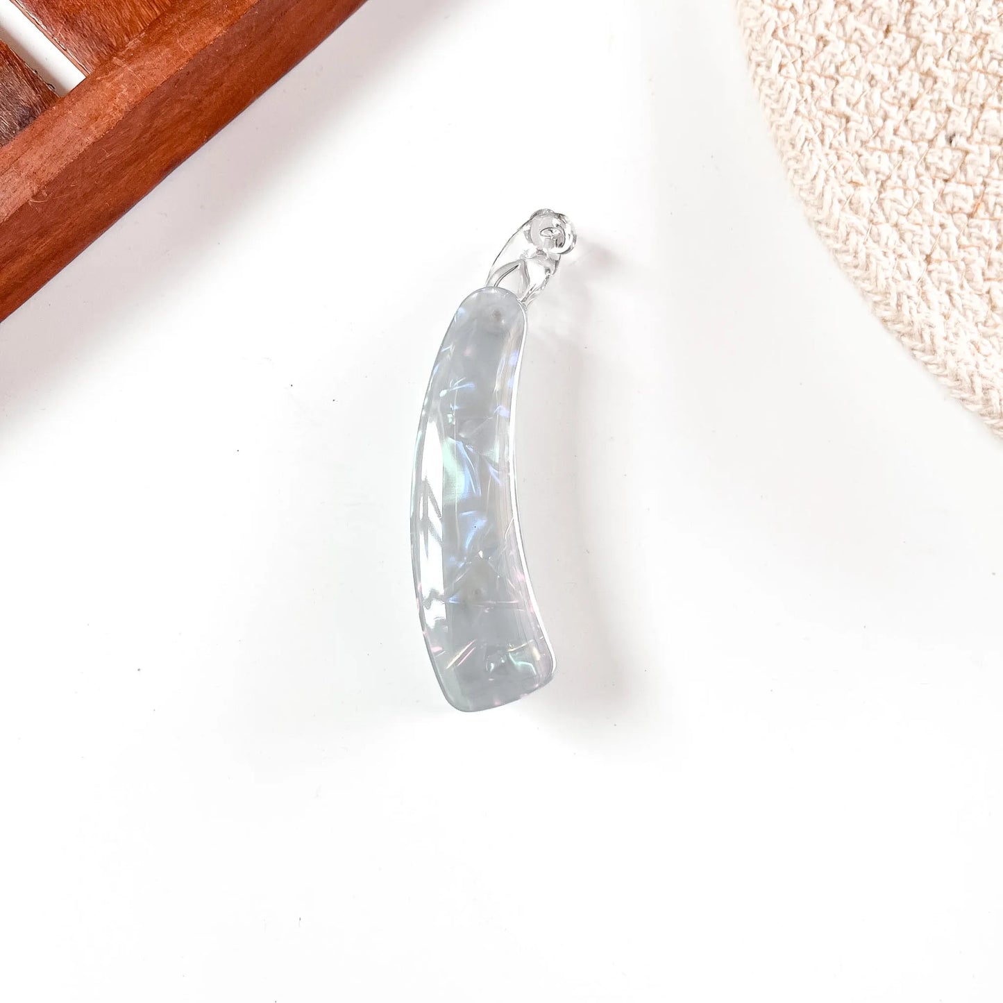 A curved, iridescent clear resin banana clip with a silver bail, resting on a white surface with decorative props in the corners.