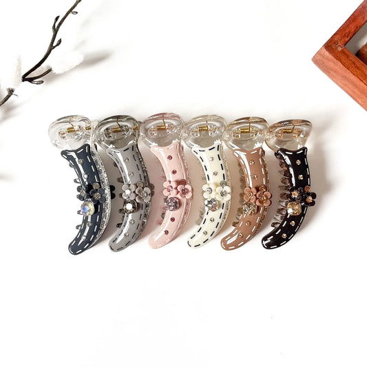 Six claw banana clips arranged in a row on a white background, each in a different color: navy, silver, light pink, cream, taupe, and black. Each clip features decorative rhinestones and faux flowers.