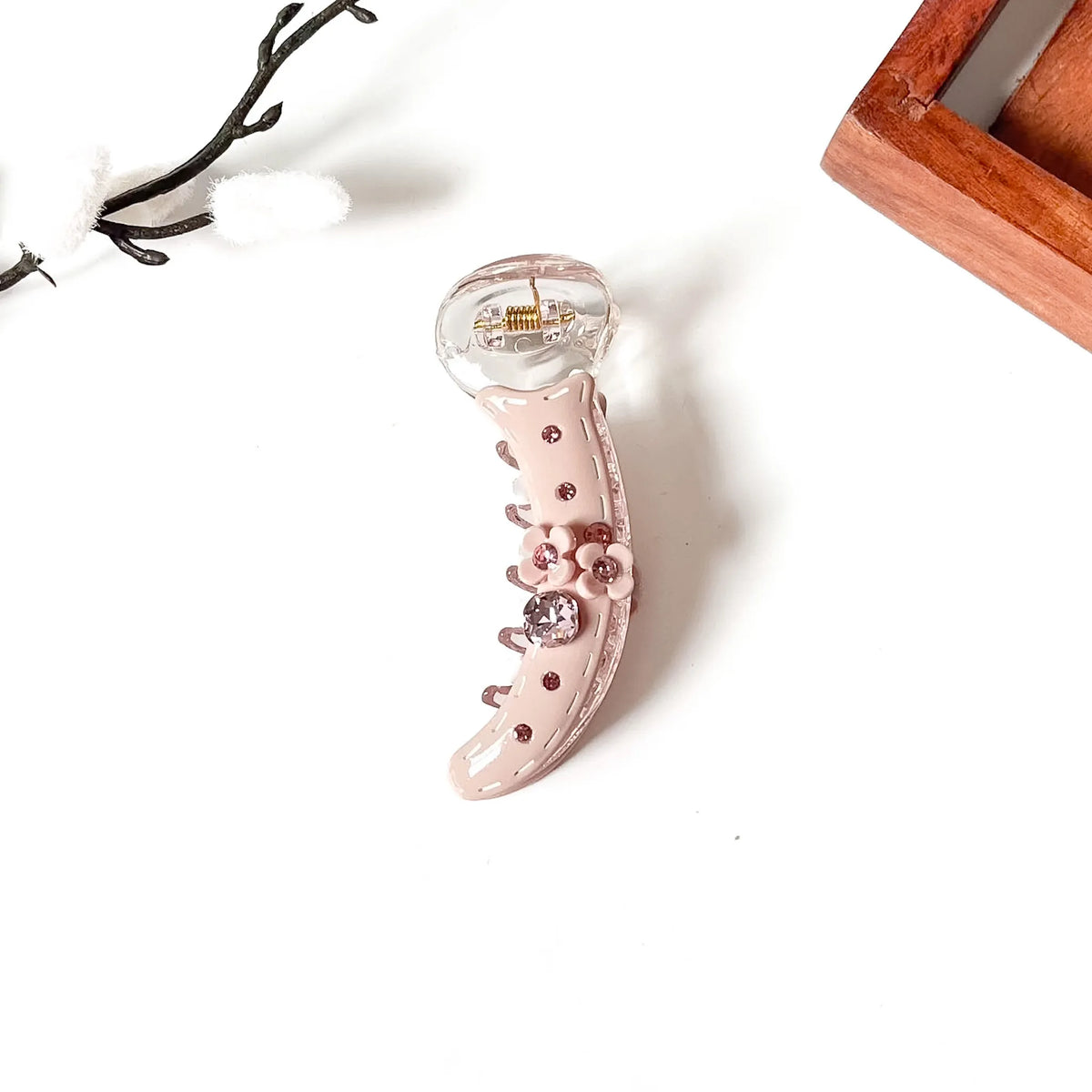 A curved claw-style banana clip in glossy pale pink with rhinestone embellishments and a clear top section, photographed on a white background with small decorative props.