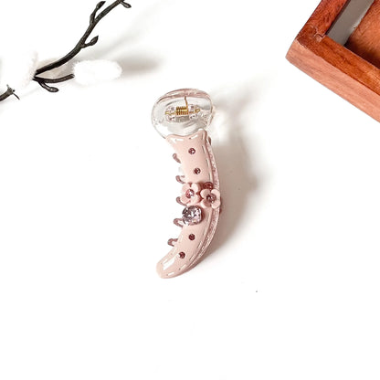 A curved claw-style banana clip in glossy pale pink with rhinestone embellishments and a clear top section, photographed on a white background with small decorative props.