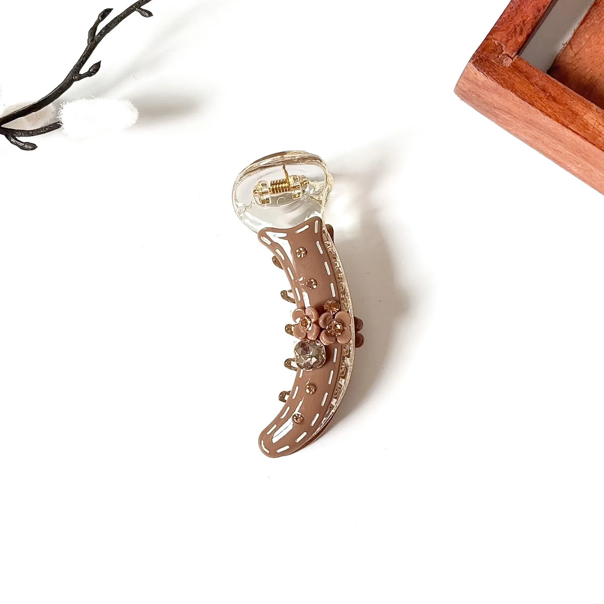 A brown claw banana clip with a clear acrylic top, gold-tone metal spring, and decorative floral embellishments, photographed on a white background with a wooden tray corner and small branch prop visible.
