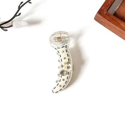 A clear acrylic claw banana clip decorated with an off-white curved body, black dashed line details, and small rhinestones, shown on a white background with decorative props.