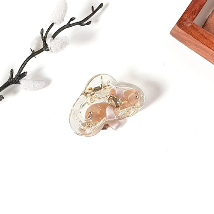 A clear acrylic hair claw clip with a light pink satin ribbon and gold-tone metal hardware, displayed on a clean white surface with decorative props.