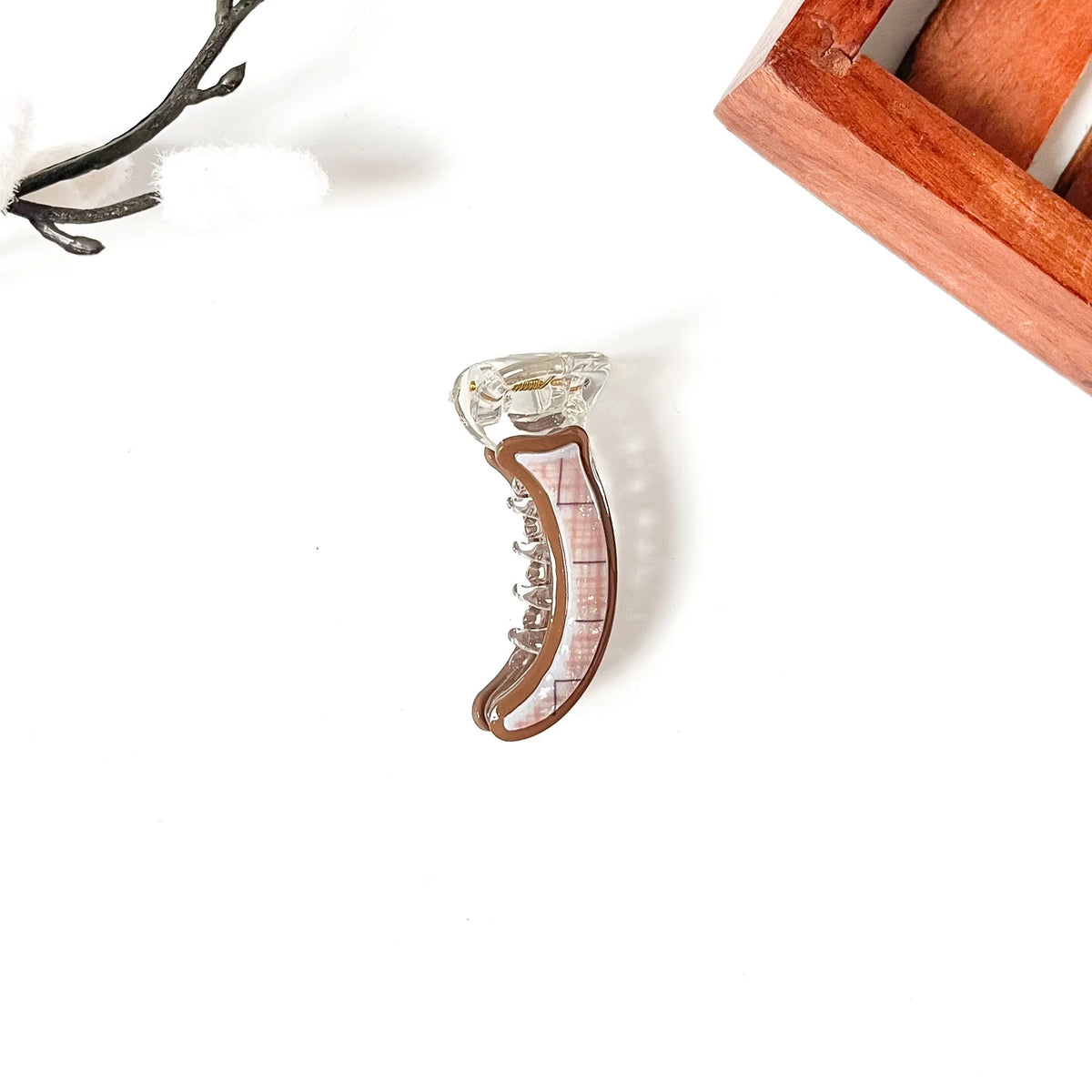 A small clear acrylic hair mini banana clip with a brown border and a pink plaid pattern, displayed on a clean white background with decorative props.