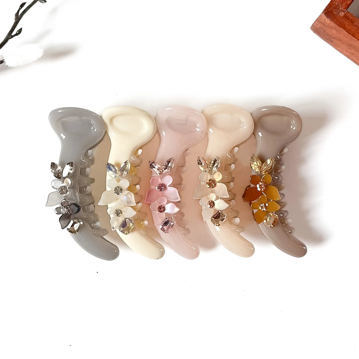 Five glossy hair banana clips arranged in a row on a white background, each decorated with small floral embellishments and rhinestone-like accents in different pastel and neutral tones.