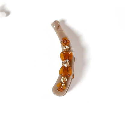 A curved beige lock banana clip decorated with a vertical row of amber and clear faceted rhinestones, shown on a plain white background.