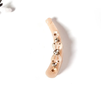 A curved, nude-beige plastic lock banana clip embellished with small metallic gold studs and three clear rhinestones, shown on a plain white background.