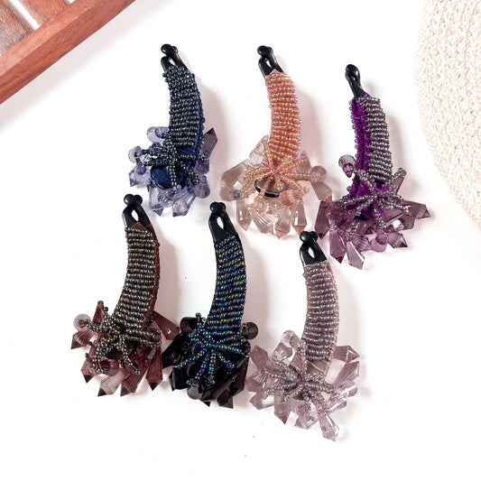 Six crystalline banana clips in black, burgundy, navy iridescent, caramel, purple, and mauve with oversized crystal gemstone clusters, arranged on cream background.