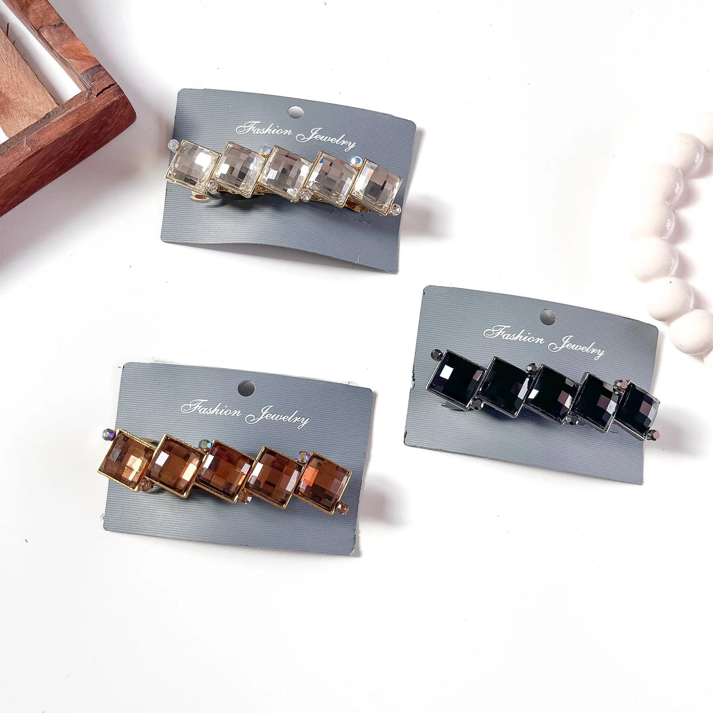 Three cube gemstone center clips in clear crystal, amber brown, and black with faceted square gemstones on branded packaging cards, displayed on cream background.