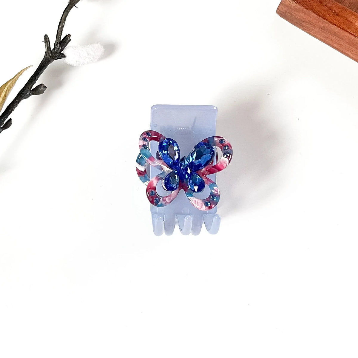 A translucent light blue hair catch clip with a decorative butterfly-shaped center featuring a marbled pattern of blue, pink, and purple, shown on a white background with decorative dried flowers.