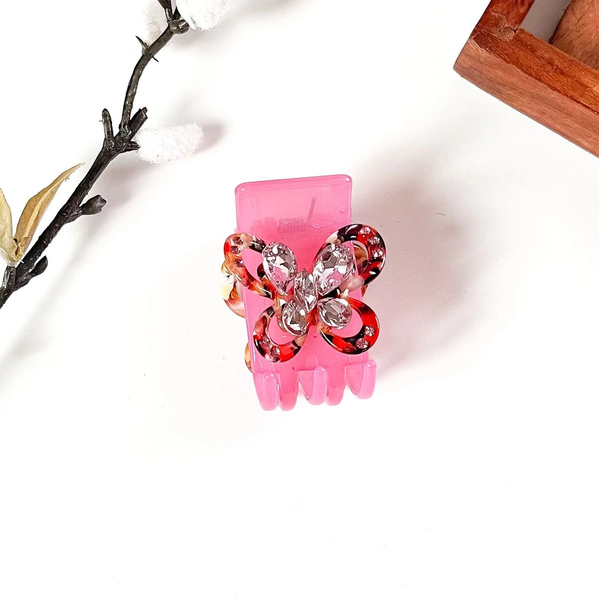 A pink hair claw catch clip decorated with a butterfly-shaped rhinestone embellishment, shown on a white background with decorative props.