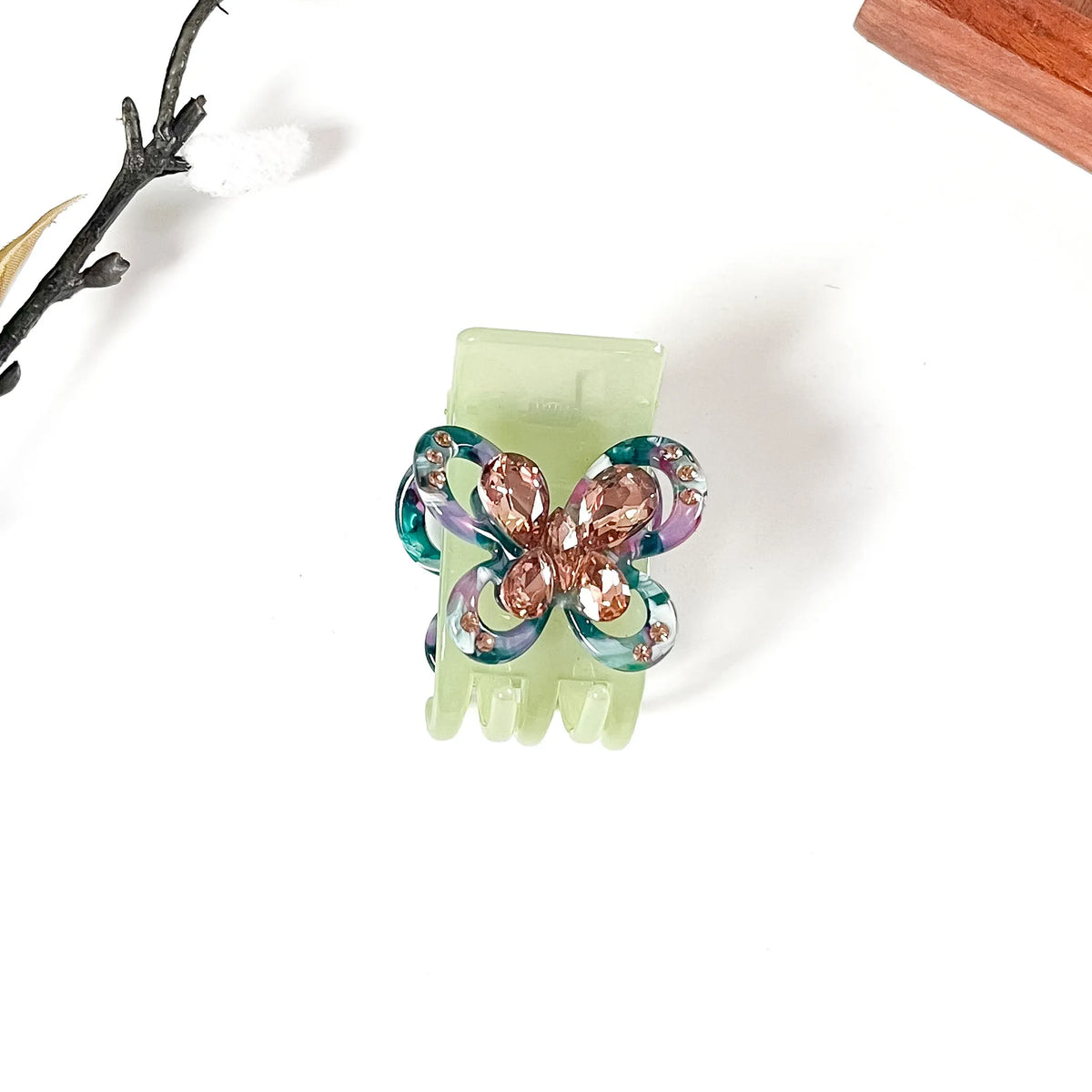 A light green claw catch clip featuring a butterfly embellishment with iridescent blue, purple, and rose gold rhinestones, shown on a white background with decorative props.