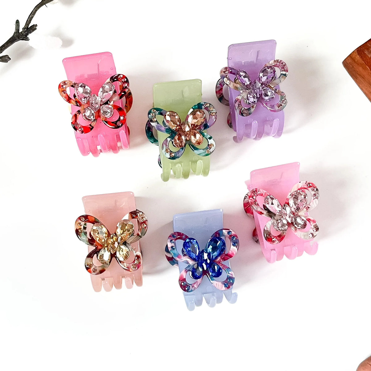 Six butterfly-shaped claw catch clips in assorted pastel and multicolor shades, decorated with rhinestone-embellished wings, arranged on a white background.