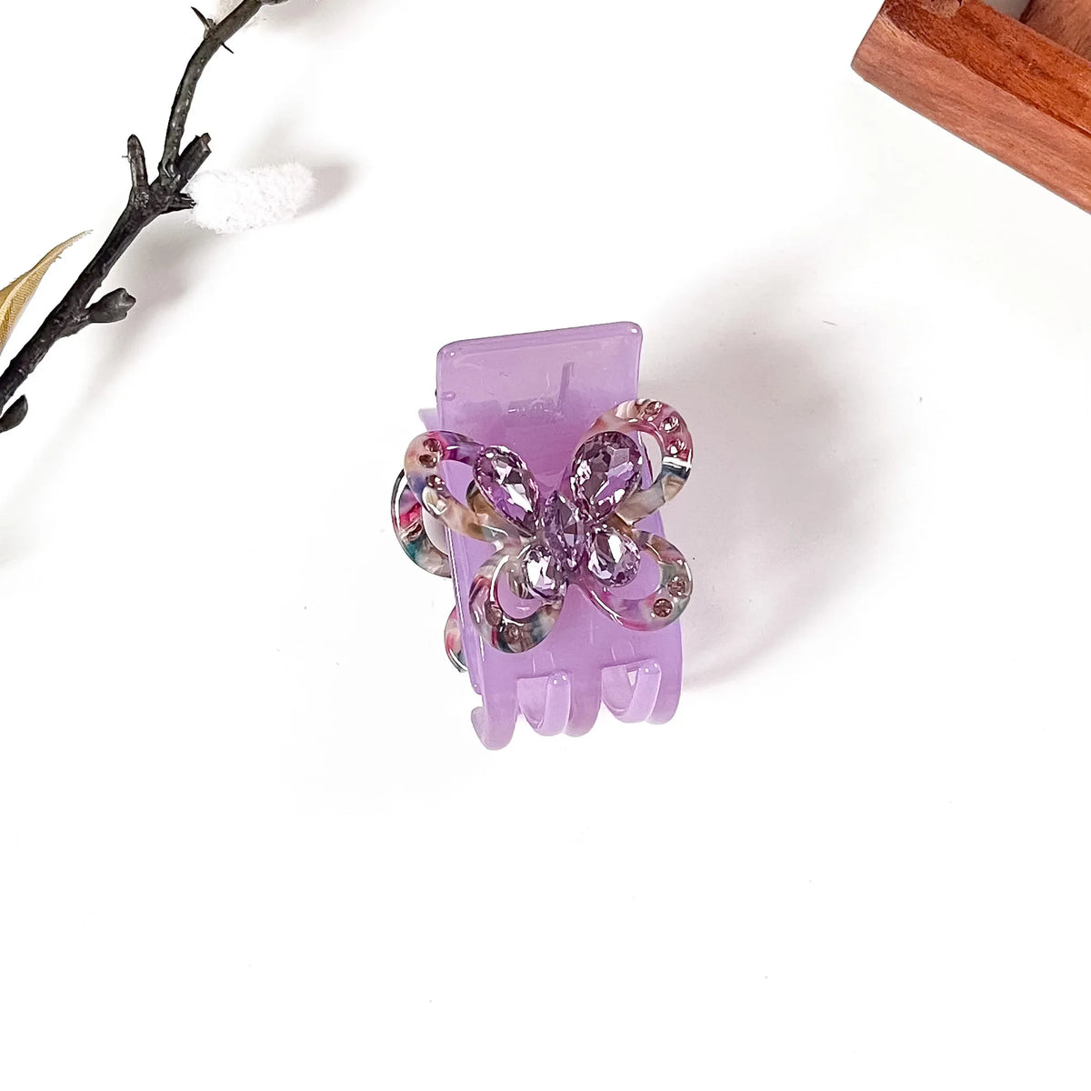 A lavender purple claw catch clip decorated with a butterfly embellishment featuring multicolor floral patterns and a central rhinestone, shown on a white background with decorative props.