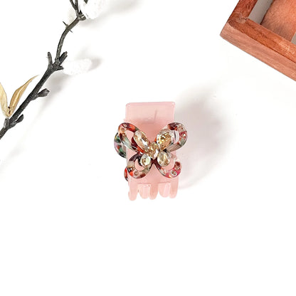 A pink claw hair catch clip decorated with a bow-shaped embellishment featuring red, green, and gold marbled swirls and clear rhinestones, displayed on a clean white background with decorative props.