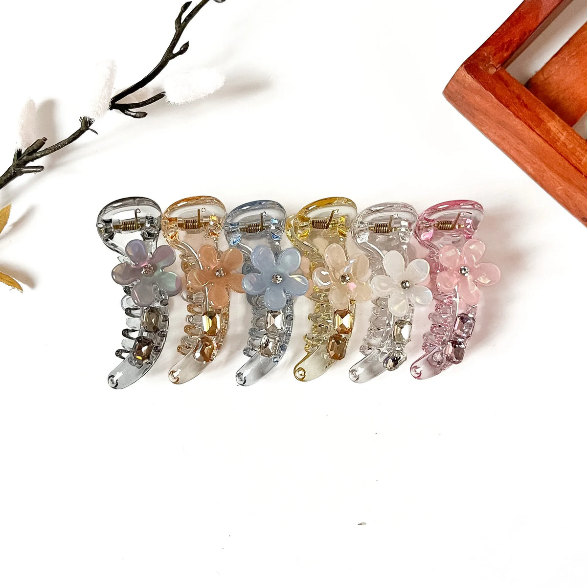Six translucent flower claw banana clips in assorted pastel and neutral colors, arranged in a row on a white background with decorative dried branches and a wooden frame corner.