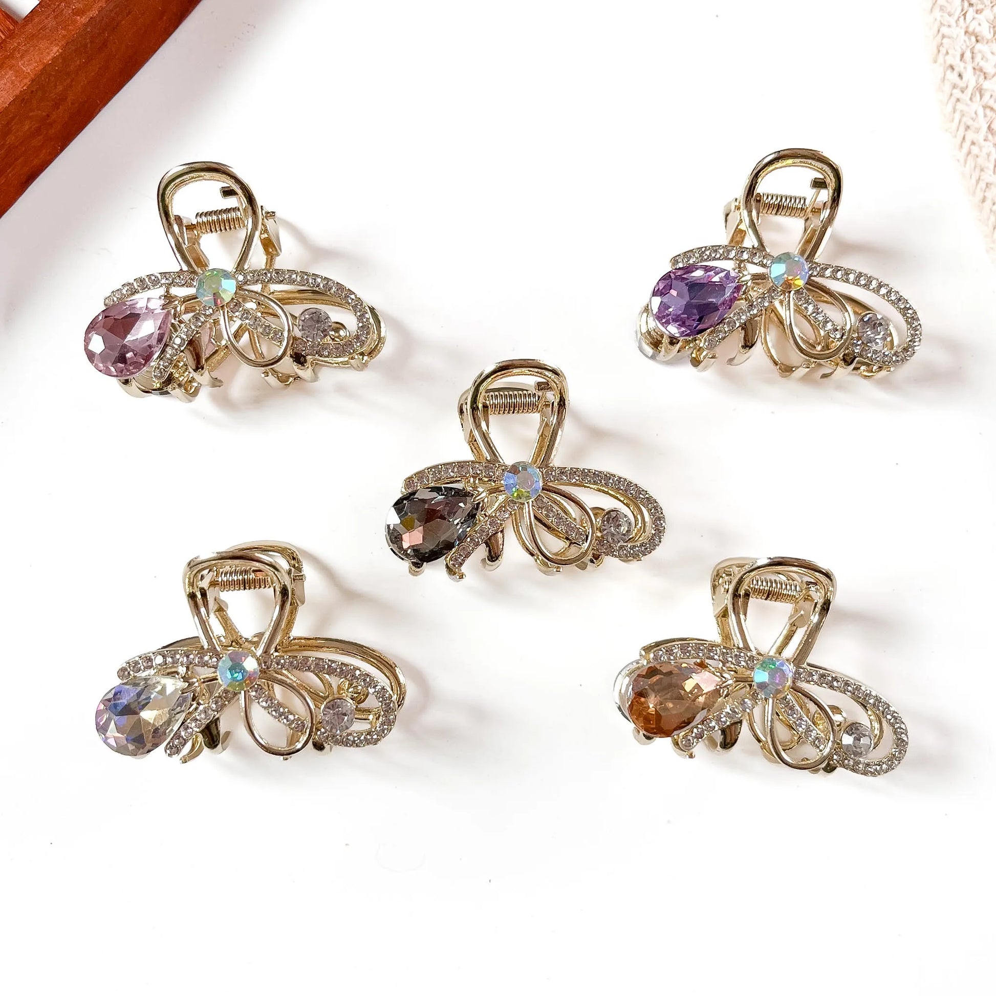 Five gemstone metal claw clips in rose pink, purple, amber, smoky, and multicolor opal with hanging faceted teardrop stones and filigree gold-tone frames, arranged flat on cream background.