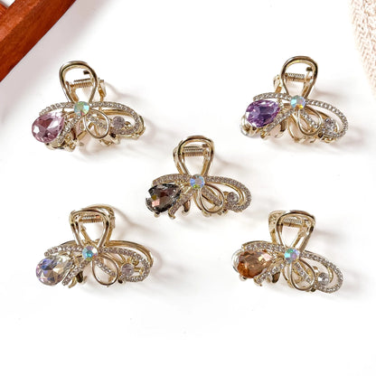 Five gemstone metal claw clips in rose pink, purple, amber, smoky, and multicolor opal with hanging faceted teardrop stones and filigree gold-tone frames, arranged flat on cream background.