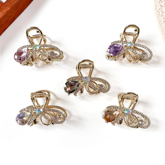 Five gemstone metal claw clips in rose pink, purple, amber, smoky, and multicolor opal with hanging faceted teardrop stones and filigree gold-tone frames, arranged flat on cream background.