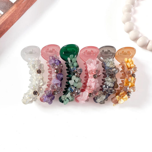 Six glam chunk banana clips in clear crystal, smoky gray, forest green, purple-pink, blush, and golden amber with oversized faceted gemstone chunks, arranged flat on cream background.