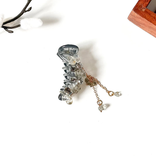 A clear resin claw banana clip embellished with clear rhinestones and dangling gold-tone chains with small clear crystal beads, photographed on a white background with decorative dried branches and a wooden corner visible.