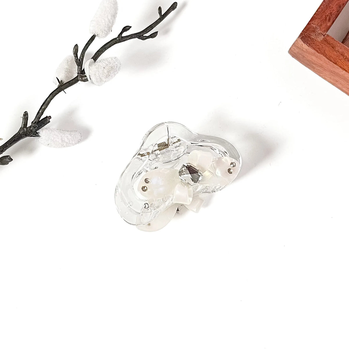 A clear resin hair claw clip with white faux pearls and a central faceted crystal, displayed on a clean white surface with decorative props.