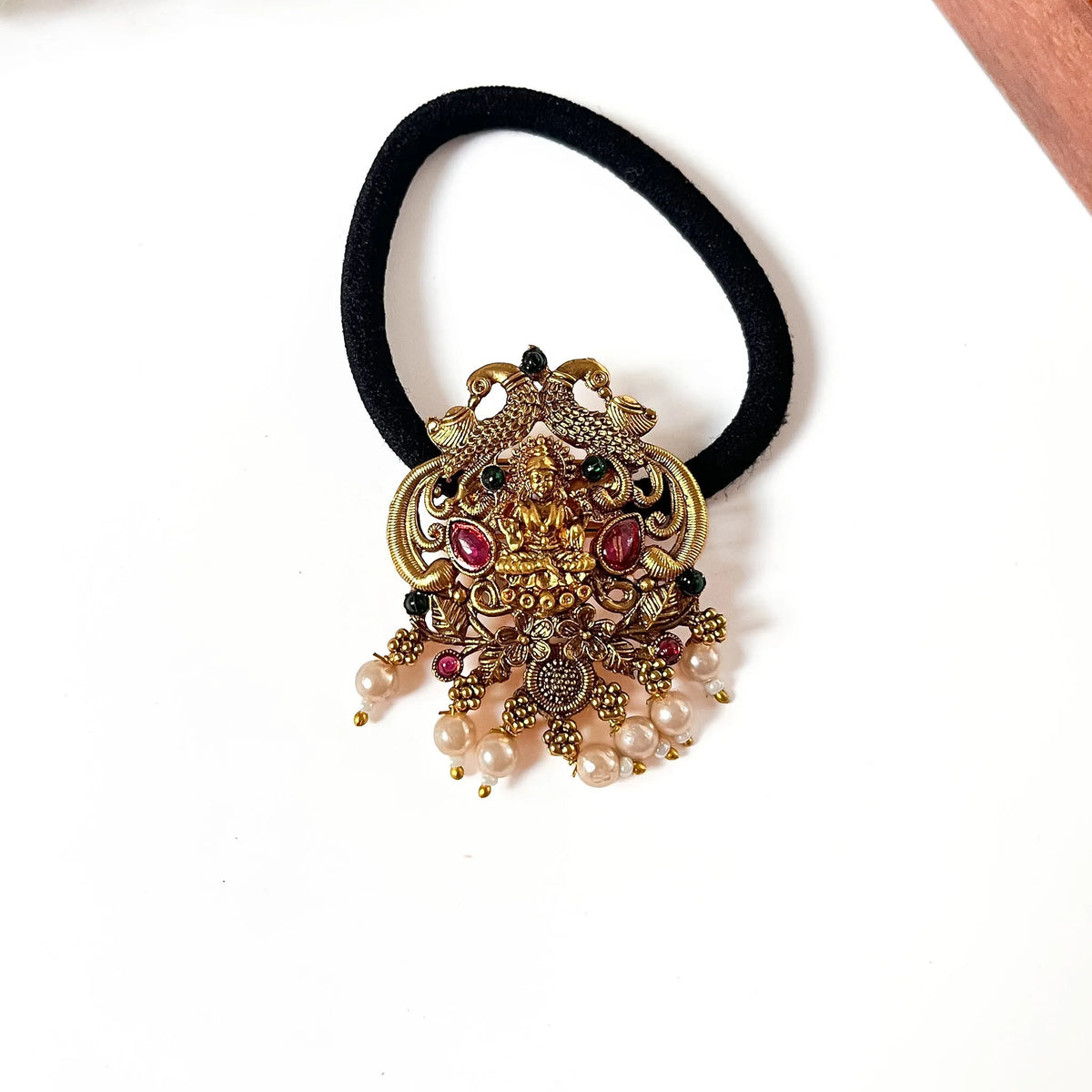 Lakshmi wing hair tie with ornate gold filigree deity charm, winged design, ruby red and black gemstones, and dangling pearl beads on black scrunchie