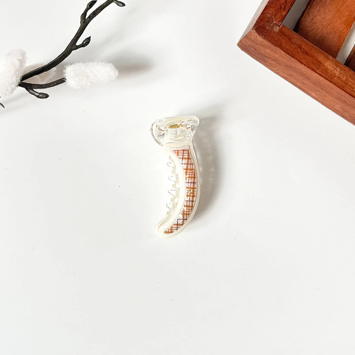 A small white crescent-shaped hair mini banana clip with a brown grid pattern, displayed on a clean white surface with decorative props.