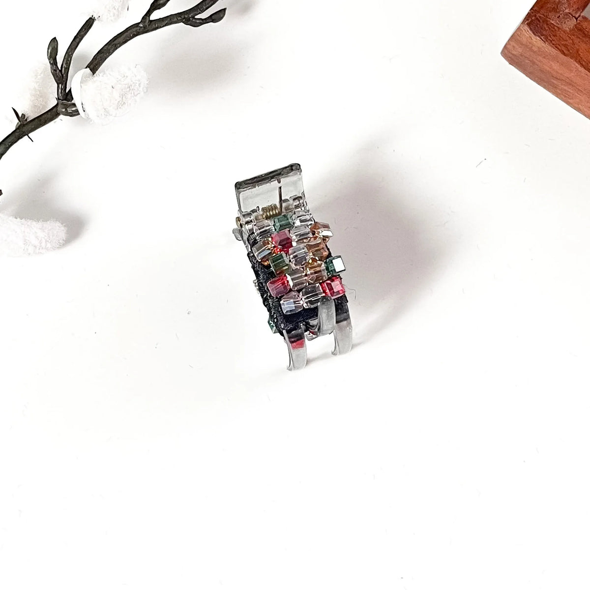 A silver-toned small catch clip decorated with multiple small colorful gemstones, photographed on a clean white surface with decorative branches in the corner.