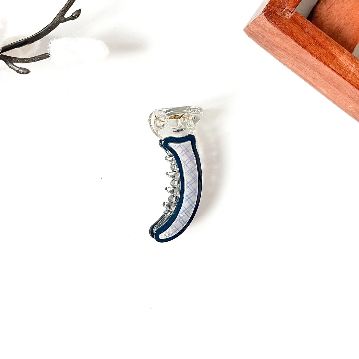 A small crescent-shaped hair mini banana clip with a navy blue border, a white and light blue grid patterned center, and clear rhinestone accents, shown on a white background with decorative props.