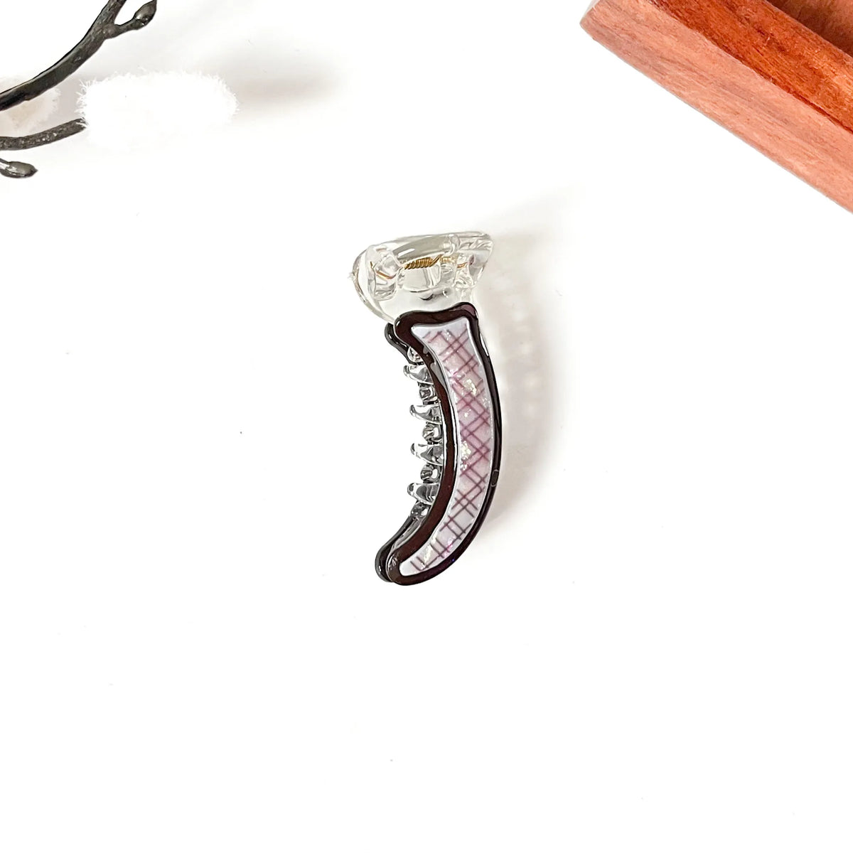 A clear plastic claw mini banana clip with a dark brown border and a pink plaid patterned insert, shown on a white background with partial wooden and metal props.