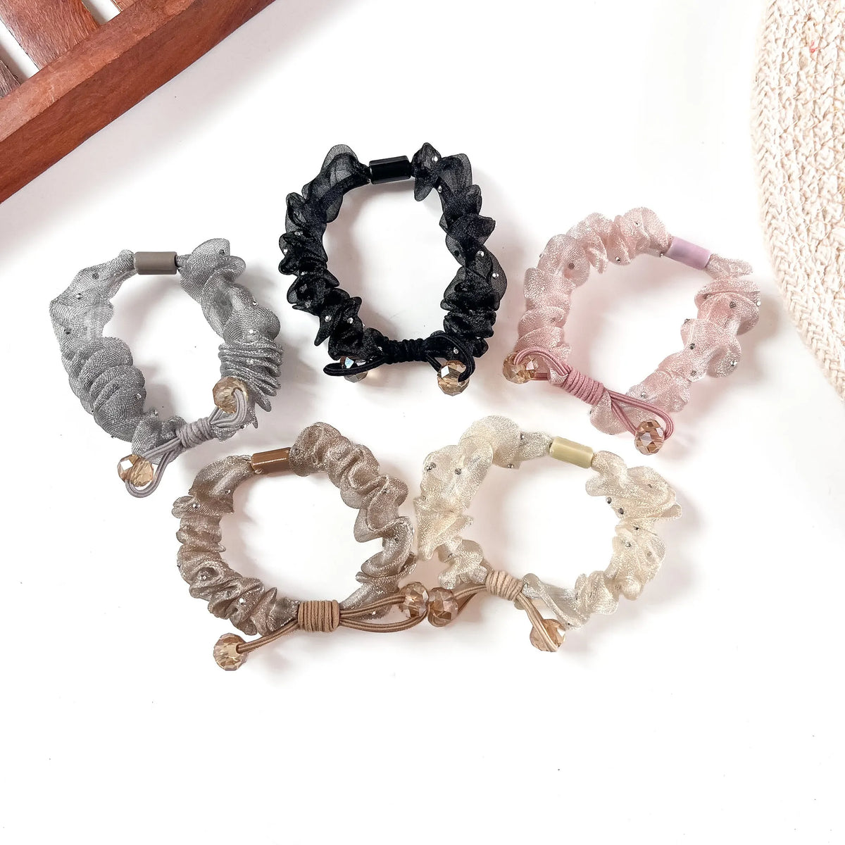 Six organza scrunchies in silver, black, pink, ivory, champagne, and rose gold, arranged on a cream linen background with wooden frame styling.