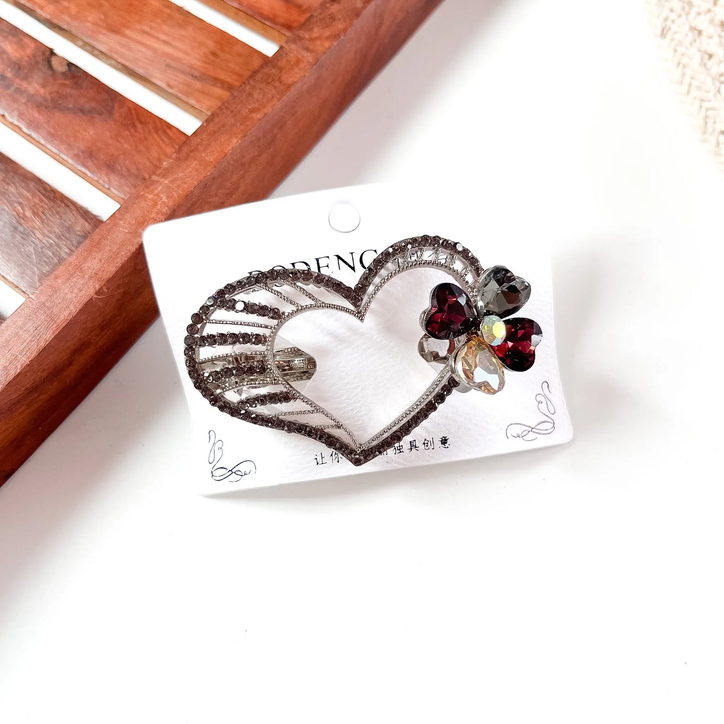 Ornate heart-shaped barrette with intricate silver filigree metalwork, oversized ruby and crystal gemstone accents, displayed on branded packaging card against cream background.