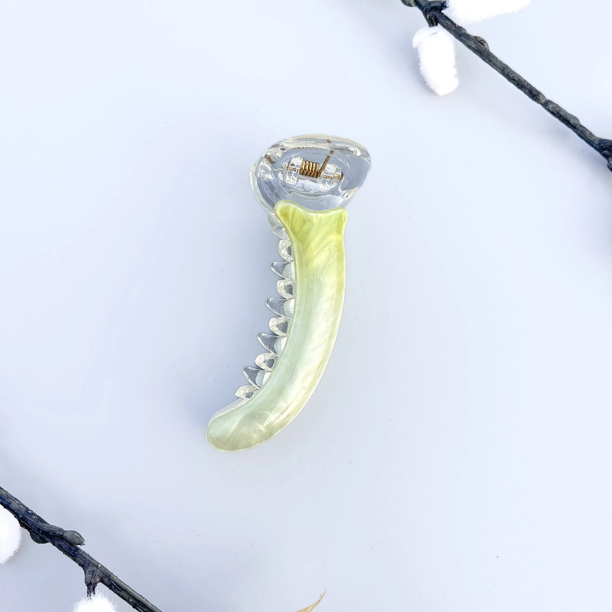 A curved banana hair clip with a translucent clear plastic body and a glossy green-yellow inner lining, photographed on a white surface with an open book and wooden object in the background.