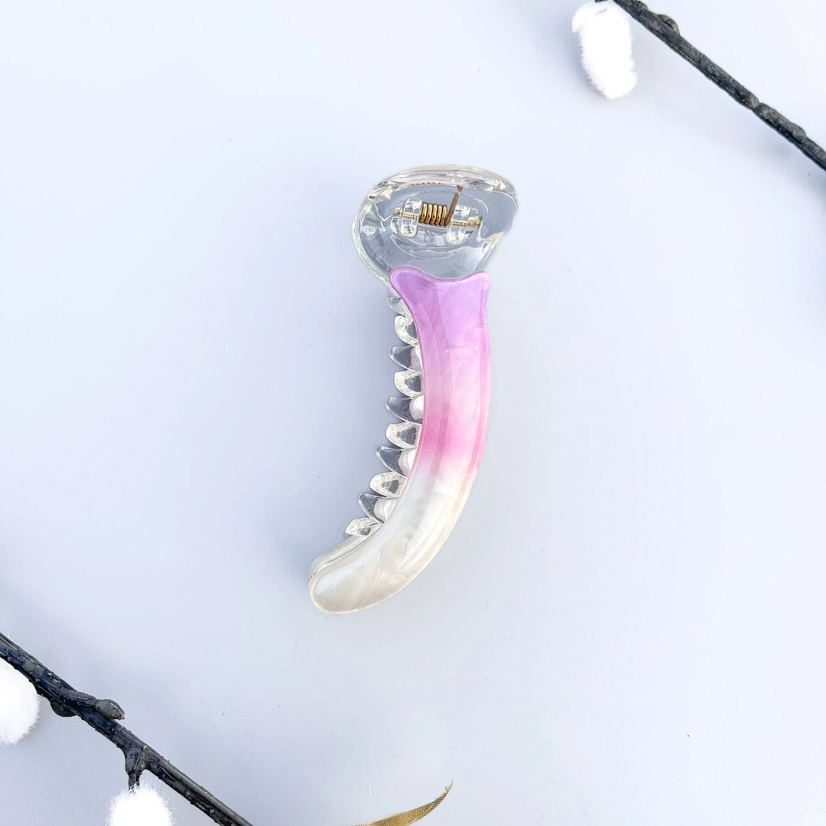 A curved banana hair clip with a translucent clear plastic body and a glossy pink inner lining, photographed on a white surface with an open book and wooden object in the background.