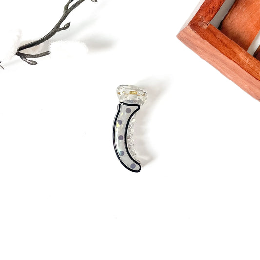 A clear acrylic claw mini banana clip with a black outer border and small gold, blue, and white polka dots, displayed on a clean white surface with decorative props.