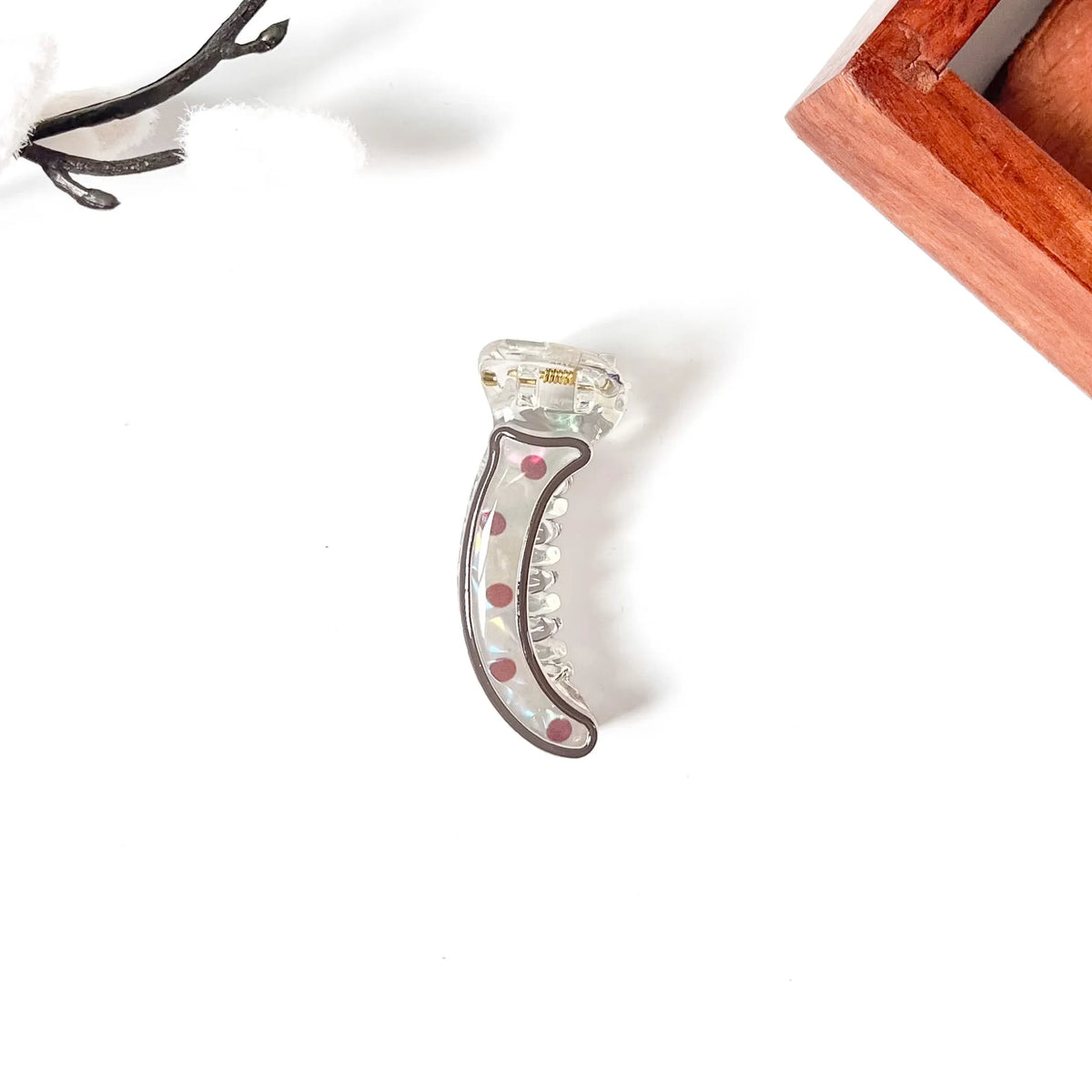 A clear acrylic crescent-shaped mini banana clip with a dark gray border and pink rhinestone accents, displayed on a clean white background with decorative props.